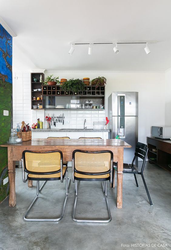 Cozinha simples, com mesa em madeira e cadeiras. Azulejos brancos, paredes brancas e chão com o piso queimado.