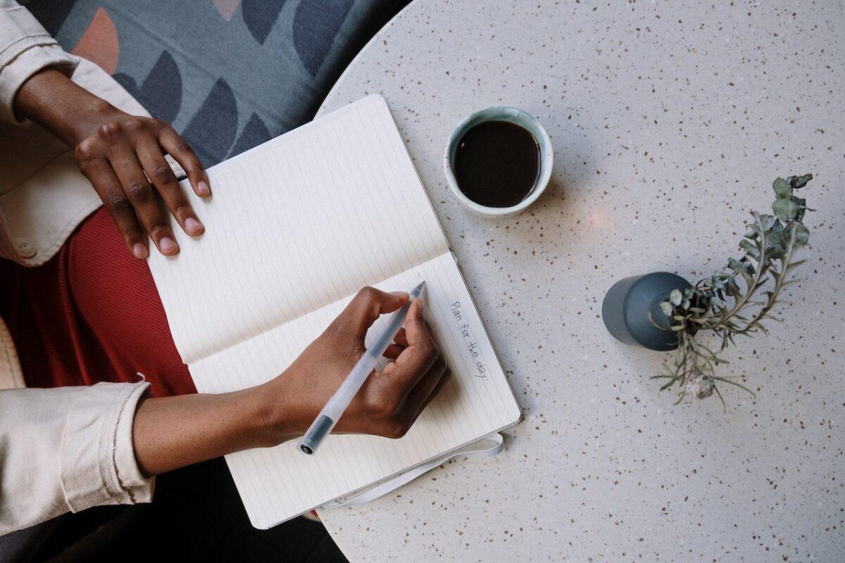 mão escrevendo em caderno em cima de uma mesa, com uma xícara de café ao lado.