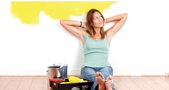 Mulher sentada ao chão com um kit de tipos de tinta, pintando uma parede branca de amarelo.