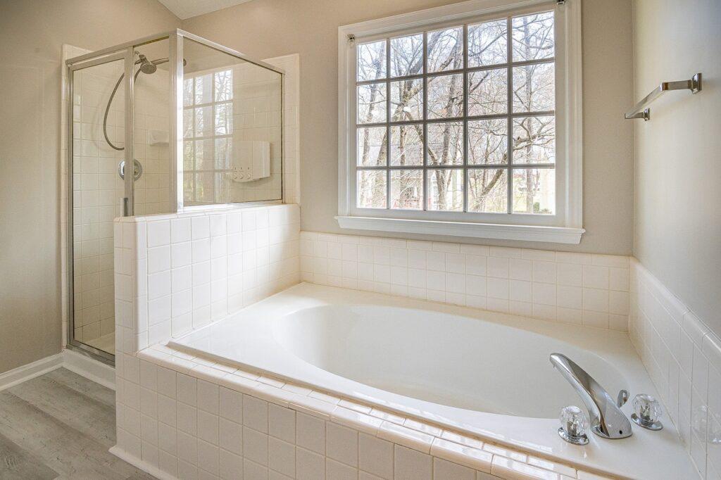 banheiro de cor clara, composto por box envidraçado e uma banheira de banho.
